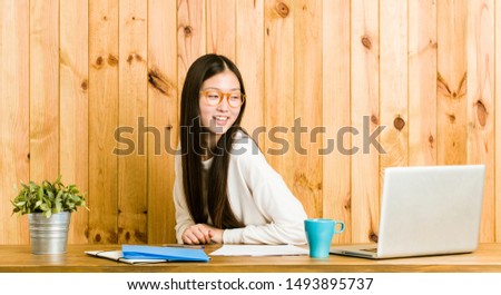 Young chinese woman studying on her desk looks aside smiling, cheerful and pleasant.