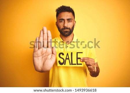 Young arab indian hispanic man holding sale banner over isolated yellow background with open hand doing stop sign with serious and confident expression, defense gesture
