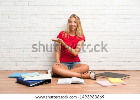 Young blonde student girl with many books on the floor extending hands to the side for inviting to come
