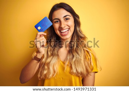Young beautiful woman holding credit card over yellow isolated background with a happy face standing and smiling with a confident smile showing teeth