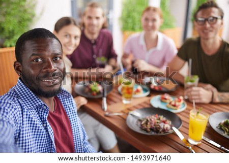 Multi-ethnic group of people posing for selfie photo while enjoying party dinner in cafe together, focus on smiling African-American man in holding camera in foreground, copy space