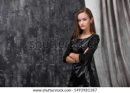 young girl in a leather dress, folded hands, stands on a gray background