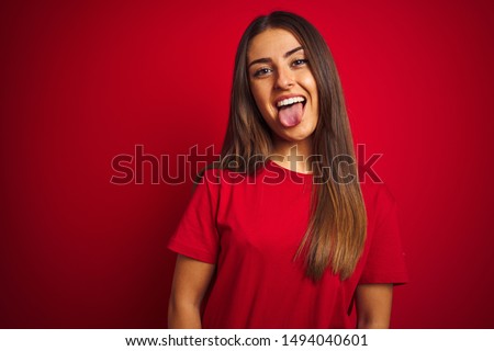 Young beautiful woman wearing t-shirt standing over isolated red background sticking tongue out happy with funny expression. Emotion concept.