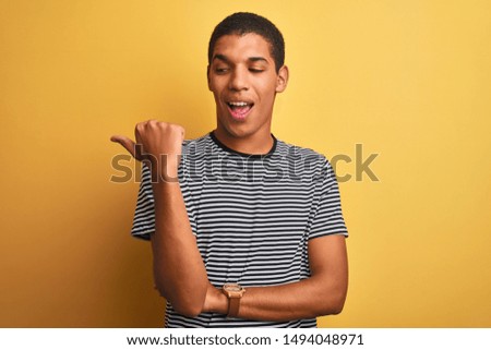 Young handsome arab man wearing navy striped t-shirt over isolated yellow background smiling with happy face looking and pointing to the side with thumb up.