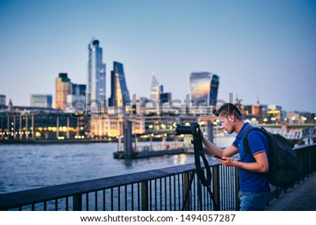 Young man photographing with tripod on embankment against urban skyline. Photographer holding smart phone with app. City life in London, United Kingdom.