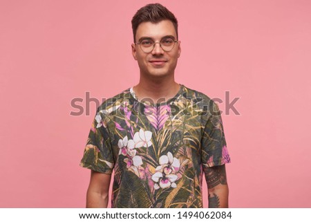 Indoor portrait of handsome young guy with tattoos wearing glasses and flowered t-shirt, smiling gently to camera, isolated over pink background