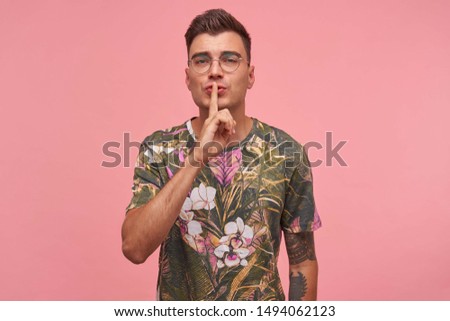 Calm pretty short haired male with index finger on his lips making hush gesture, wearing glasses and flowered t-shirt, standing over pink background