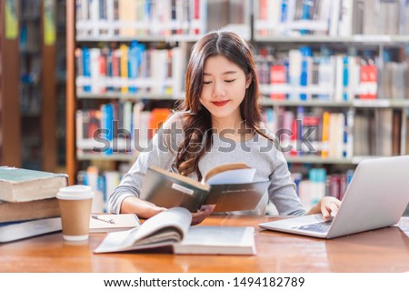Asian young Student in casual suit doing homework and using technology laptop in library of university or colleage with various book and stationary over the book shelf background, Back to school Royalty-Free Stock Photo #1494182789