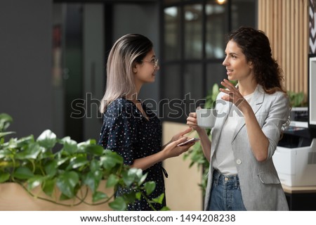 Two young diverse business women chatting during work break standing in modern office, caucasian female employee share ideas talking with asian colleague discussing team project at corporate meeting Royalty-Free Stock Photo #1494208238
