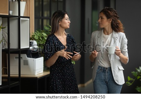 Two serious diverse female colleagues team talk walk in modern office, asian businesswoman having business conversation with caucasian coworker discuss project work meeting in company hallway space Royalty-Free Stock Photo #1494208250