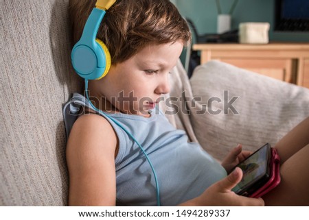 4 years boy in headphones watching videos with smartphone. Enjoying sitting on sofa in living room at home