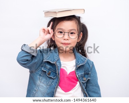 Asian little cute girl wearing glasses and put the book on head. Preschool lovely kid with the book on her head and pointing finger up. Learning and education of kid. Royalty-Free Stock Photo #1494361640