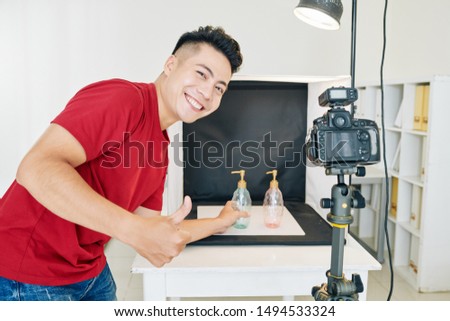 Cheerful Vietnamese photographer showing thumbs-up when putting soap dispensers against black background in studio