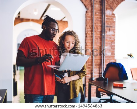 Casual African American man and young curly woman watching laptop together while standing in contemporary light study room of university