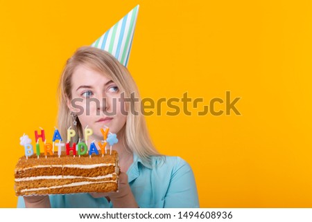 Crazy cheerful young woman in paper congratulatory hat holding cakes happy birthday standing on a yellow background with copy space. Jubilee congratulations concept.