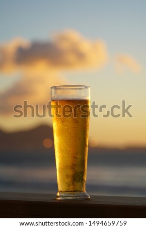 a glass of beer standing in the center of the picture with the ocean and mountains blurred in the background a evening sky in summer                              