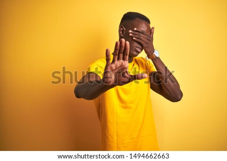 Young african american man wearing casual t-shirt standing over isolated yellow background covering eyes with hands and doing stop gesture with sad and fear expression. Embarrassed and negative