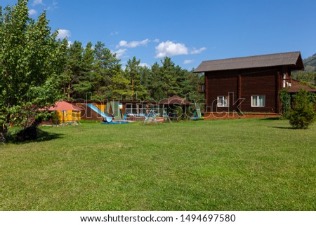 Two-storey house made of timber, surrounded by a green lawn, flowers and an alley, next to a children's play area with a pool, a slide and various swings in the background high mountains with blue sky