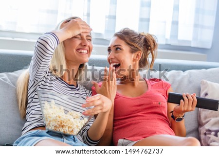 Portrait of funny and happy young women watching comedy in bed and laughing. Cheerful friends eating tasty popcorn and looking movie with gladness. Cozy and friendly atmosphere