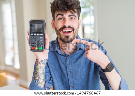 Young man holding dataphone point of sale as payment very happy pointing with hand and finger