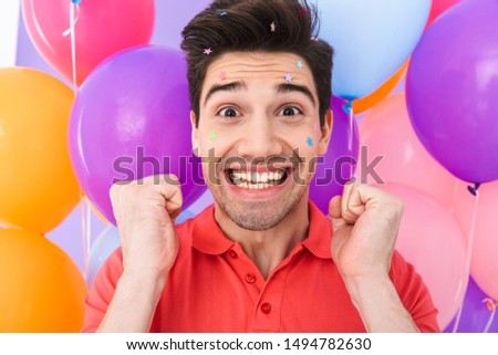 Image closeup of delighted brunette man rejoicing with glitter stars on his face and multicolored air balloons isolated over purple background
