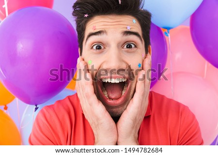 Image closeup of cheerful brunette man rejoicing with glitter stars on his face and multicolored air balloons isolated over purple background