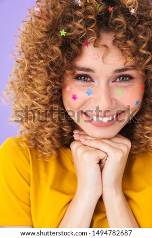 Image closeup of young pretty curly girl having fun and smiling with multicolored glitter stars on her face isolated over violet background