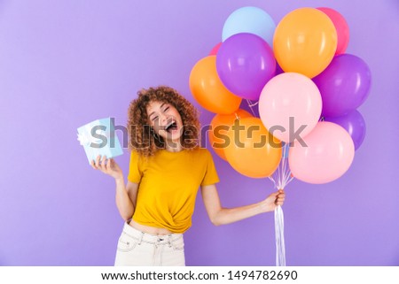 Image of beautiful young woman celebrating birthday with multicolored air balloons and present box isolated over violet background