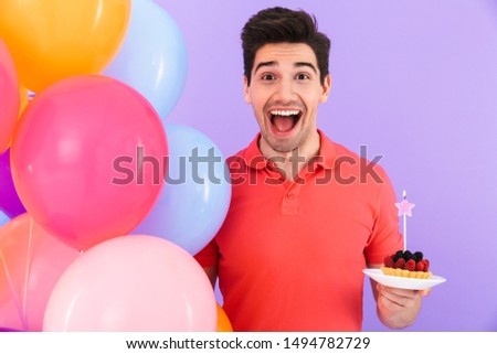 Image of optimistic joyful man celebrating birthday with multicolored air balloons and piece of pie isolated over violet background