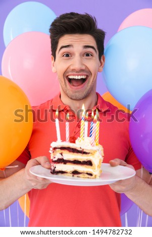 Image of handsome joyful man celebrating birthday with multicolored air balloons and piece of cake isolated over violet background
