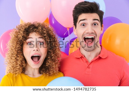 Image of delighted couple man and woman laughing at camera while standing with multicolored air balloons isolated over violet background