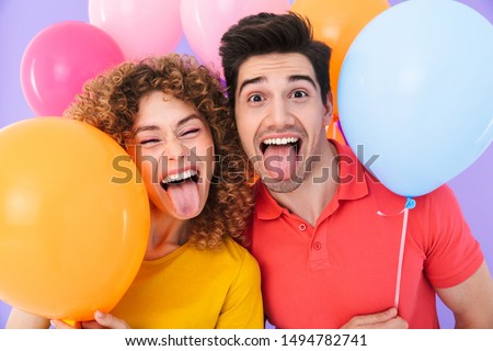 Image of cheerful couple laughing and sticking out tongues at camera while standing with multicolored air balloons isolated over violet background