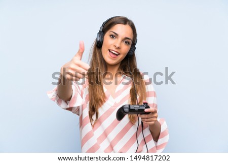 Young woman playing with a video game controller over isolated blue wall with thumbs up because something good has happened