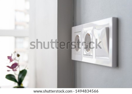 A closeup view of a group of white european electrical outlets and a switch located on a gray wall in a light modern kitchen by the window. Selective focus. Blurred background Royalty-Free Stock Photo #1494834566