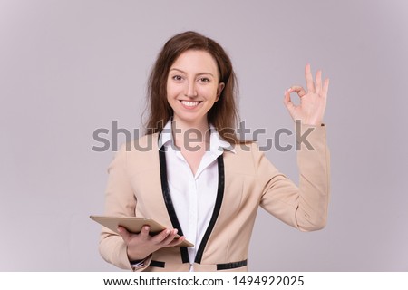 Cheerful millennial woman in an elegant blazer shows OK holding a tablet in her hands posing on a pale purple background. Successful woman advertises office application. Advertising space