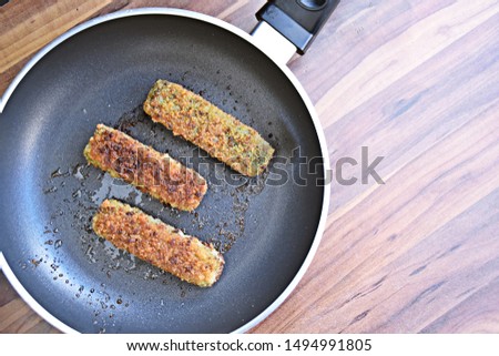 Picture from the top of a pan with fried fish fingers with spinach on a dark wooden surface - Delicious freshly fried dark fish sticks in a black teflon pan