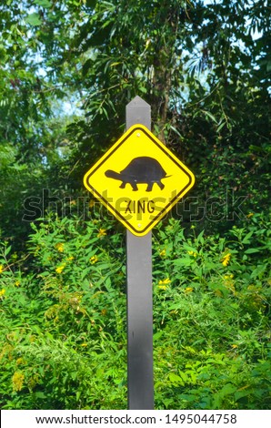 close up of yellow turtle crossing sign with black turtle icon with greenery background outdoors on a brown pained wood pole
