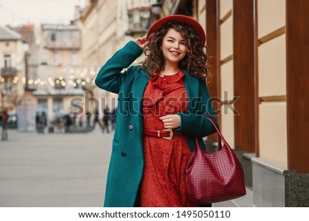 Happy smiling fashionable curvy woman wearing trendy autumn outfit: orange hat, snakeskin print dress, belt, green coat, holding red wicker leather bag, posing in street of European city. Copy space Royalty-Free Stock Photo #1495050110