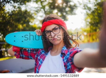 Selfie portrait of funny young woman in glasses and red plaid shirt. Hipster girl fooling around in skatepark while taking picture with skateboard in skatepark at sunny bright day. 