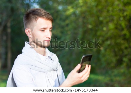 A sad young man is standing in the park, holding a smartphone in his hands, receives an unexpected message, a shocked, offended guy, a male look at the mobile phone, is stunned by the offer.
