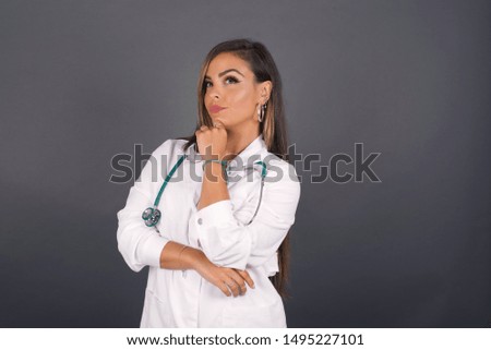 Portrait of thoughtful smiling doctor woman keeps hand under chin, looks sideways, thinking about something with interest, dressed casually, poses against gray studio. Taking decisions concept.