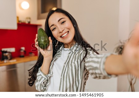 Asian woman smiles, holds avocado and takes selfies in kitchen
