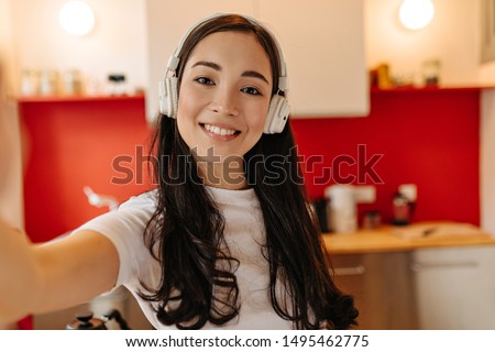 Brown-eyed woman in white headphones smiles and takes selfie in kitchen