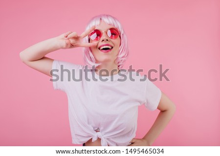 Ecstatic caucasian girl in trendy white t-shirt posing with peace sign and laughing. Indoor photo of dreamy european woman in shiny peruke and sunglasses.