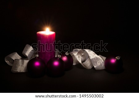 Arrangement of beautiful illuminated purple colored Christmas balls with a silver and white colored ornate ribbon and a burning candle on black background