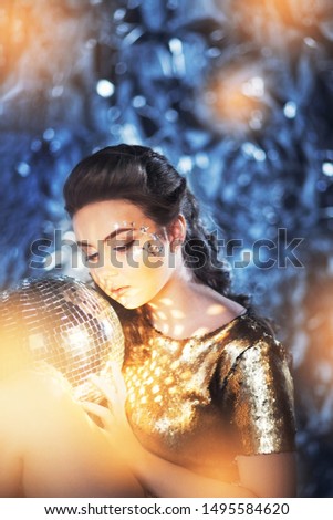 Beautiful young girl in the studio with a shiny blue background with a disco ball from the fragments of a mirror in her hands, stylization of the 70s, magic, New Year, winter