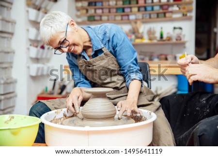 Senior woman spinning clay on a wheel with a help of a teacher at pottery class