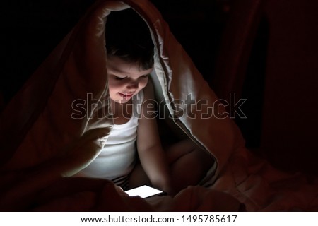 Cute little boy watching a movie at night, sitting down on bed in the bedroom with mobile. Kid watching cartoons on daddy's smartphone before he goes to sleep in night. Technology concept. 