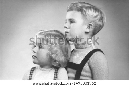 Early 1950s duo portrait of a young boy and girl with blond hair and curls looking sideways.
