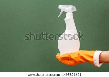 Woman's hand in orange protective glove with plastic bottle of glass and tile cleaner on the green background. Washing and cleaning concept.